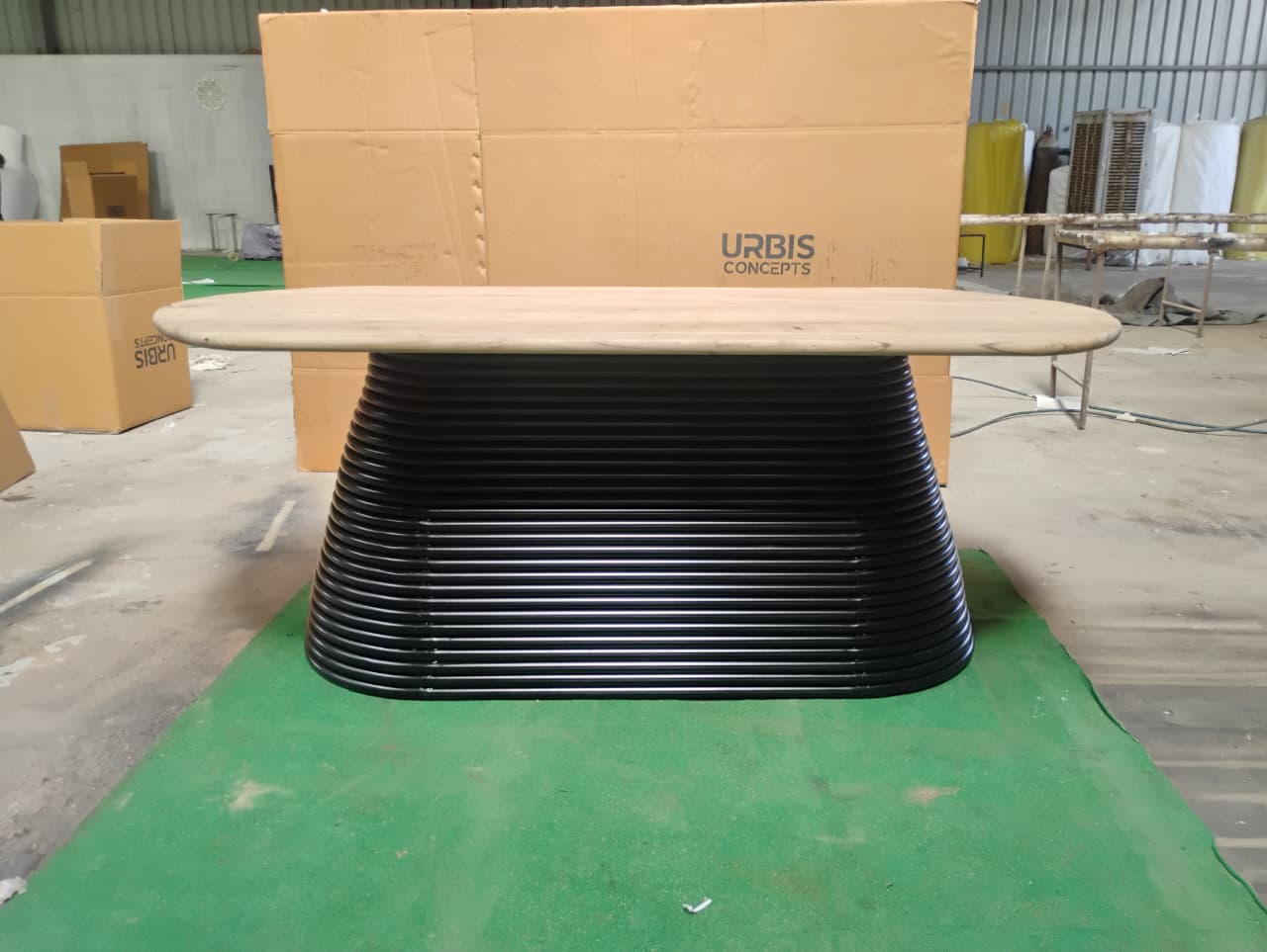 Round black base with a wooden top on a green surface, with 'Urbis' branding in the background.